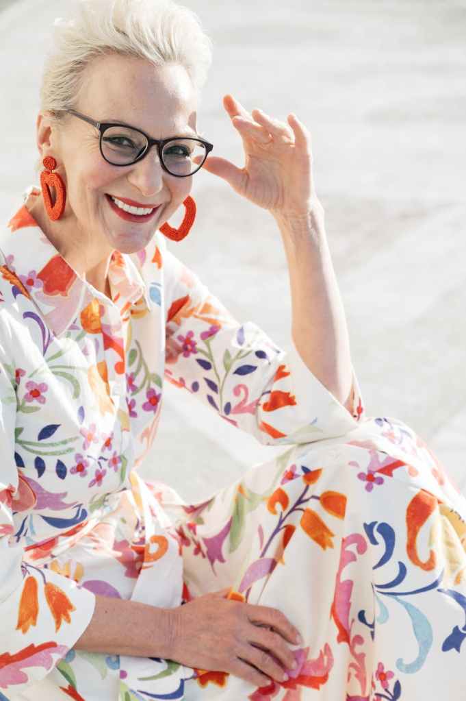 Stylish blonde woman aged fifties wearing glasses, orange earrings, colourful floral print  dress white background, orange pink and blue print