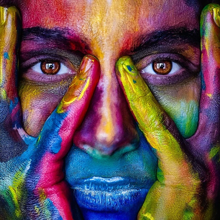 Striking Woman holding her face in her hands, eyes open covered in multicolour paint and blue lips