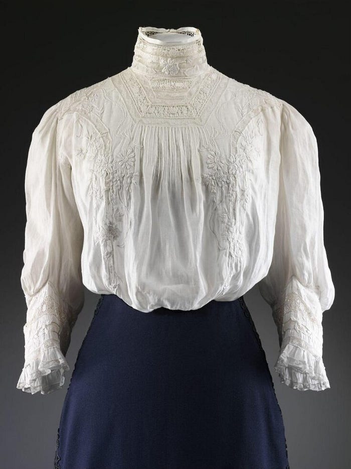 High-necked blouse of fine white lawn with full gathered elbow-length sleeves and a high band collar.